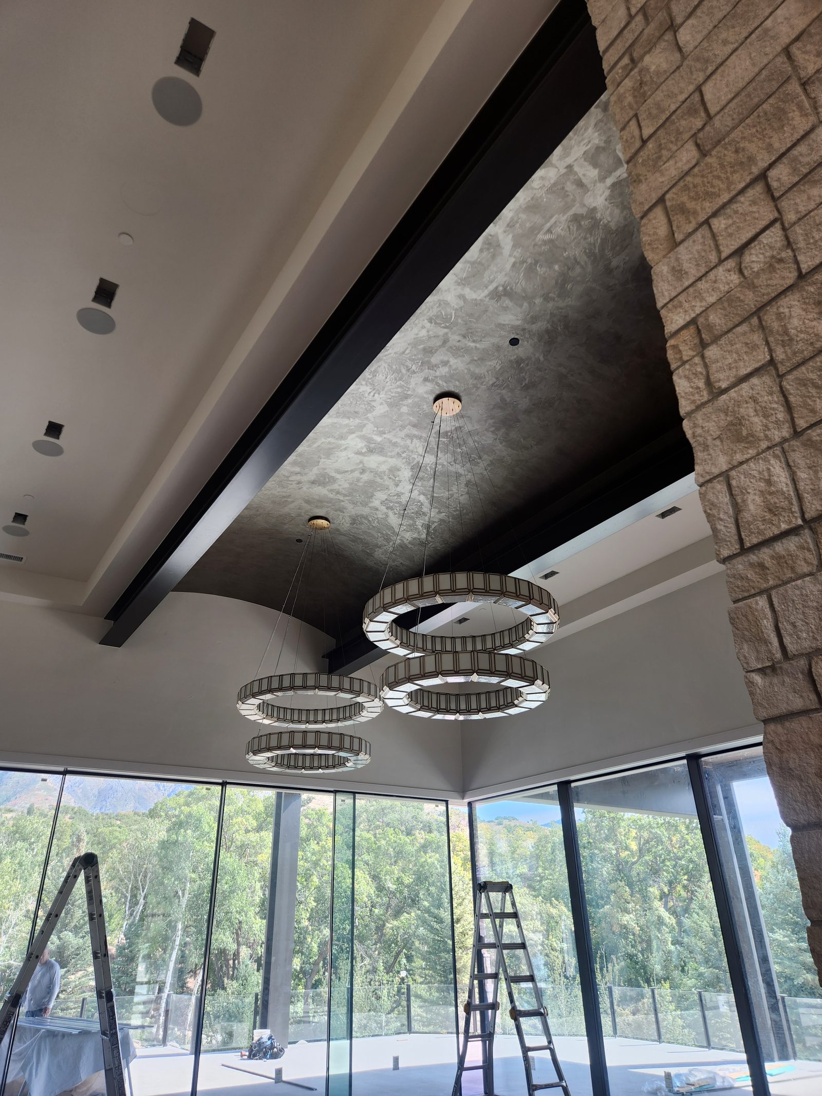 Custom-home great room under construction with tiered chandeliers and a stone fireplace, photographed during the Canvas Drywall finishing phase
